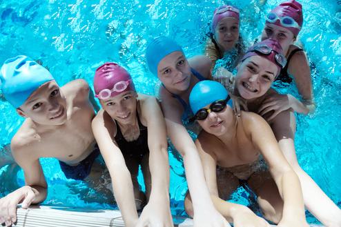 Gruppennahaufnahme von sieben Schwimmern im Wasser, lächelnd, mit Schwimmkappen und Brillen, vor einem blauen Hintergrund.
