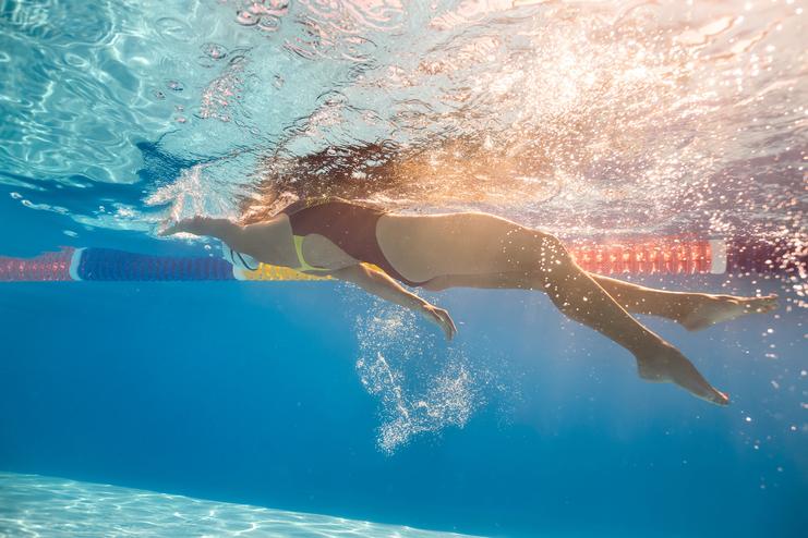 Unterwasseraufnahme einer Schwimmerin in einem Bikini, die kräftig durch das Wasser stößt, mit bunten Schwimmbahnmarkierungen.